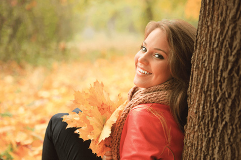 a woman outside amongst autumn leaves leaning against a tree and smiling