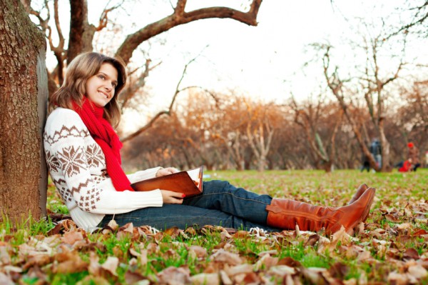 a woman wearing a jumper, scarf, dark blue jeans and brown calf boots leaning against a tree and autumn leaves scattered around her